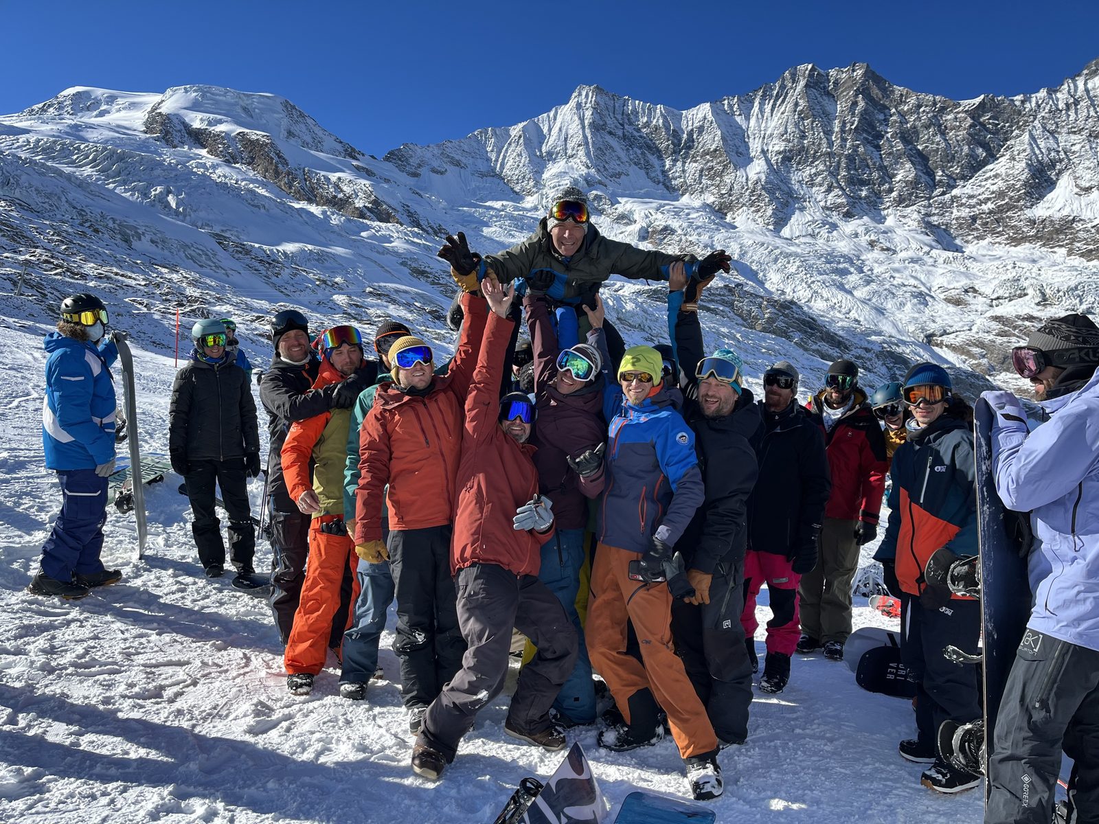 Eine grosse Gruppe Snowboarderinnen und Snowboarder hebt Roland vor vergletscherten Bergen in die Luft