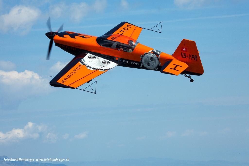 Die orange Votec 322 mit Kennung HB-YPR im Flug vor blauem Himmel